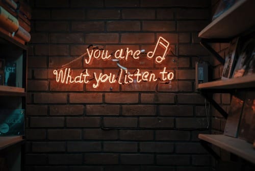 Brick wall pictured with neon sign "you are what you listen to" glowing with music note; bookshelves adjacent.